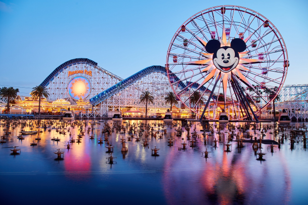 Eine lebendige Dämmerungsszene im Disneyland California Adventure mit einem riesigen Mickey-Maus-Rad im Vordergrund, einer Achterbahn im Hintergrund und Booten auf dem Wasser, mit einem Himmel aus Orangen, Pink- und Purpurfarben und silhouettenartigen Bäumen.