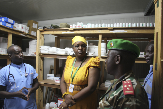 Vier Personen in einer Apotheke, mit einer Frau in einem gelben Kleid und grüner Mütze in der Mitte, flankiert von zwei Männern in blauen Hemden mit Stethoskopen, umgeben von Medikamentenflaschen und -schachteln auf der Theke und Regalen.