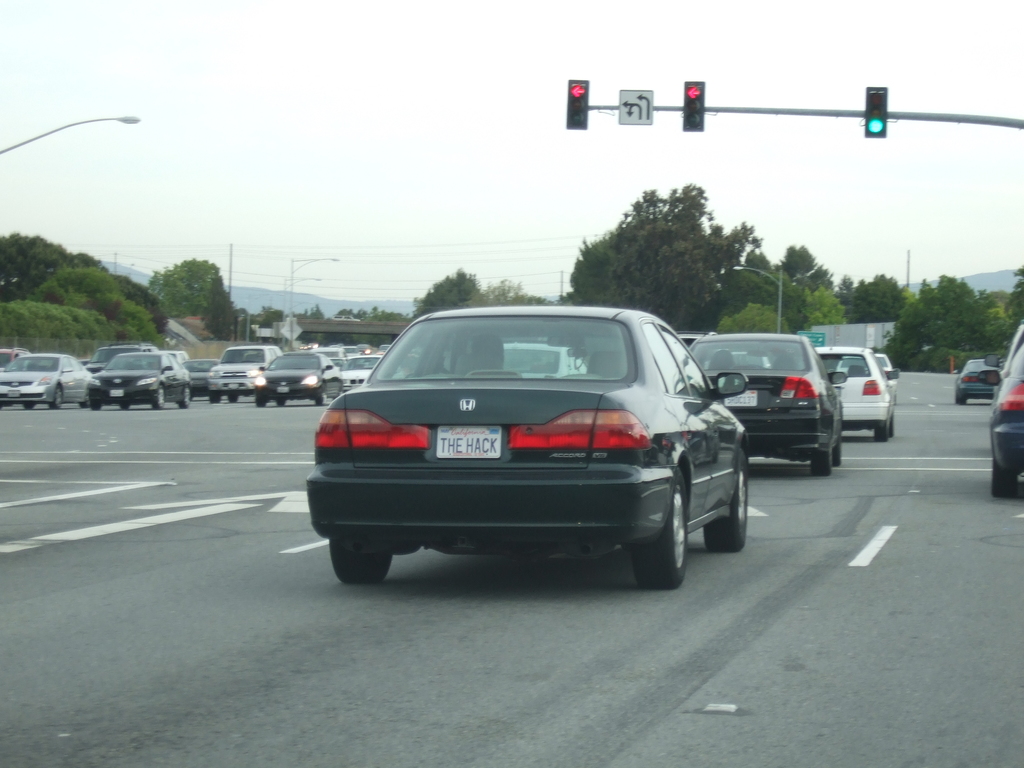 Eine vielbefahrene Straße mit mehreren Fahrzeugen, drei Strommasten und einer Ampel auf der rechten Seite unter einem klaren Himmel.
