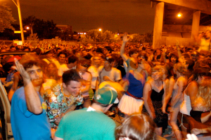 Menschen tanzen vorne bei einem Konzert mit Lichtern, Masten, B├Ąumen und anderen Objekten im Hintergrund.