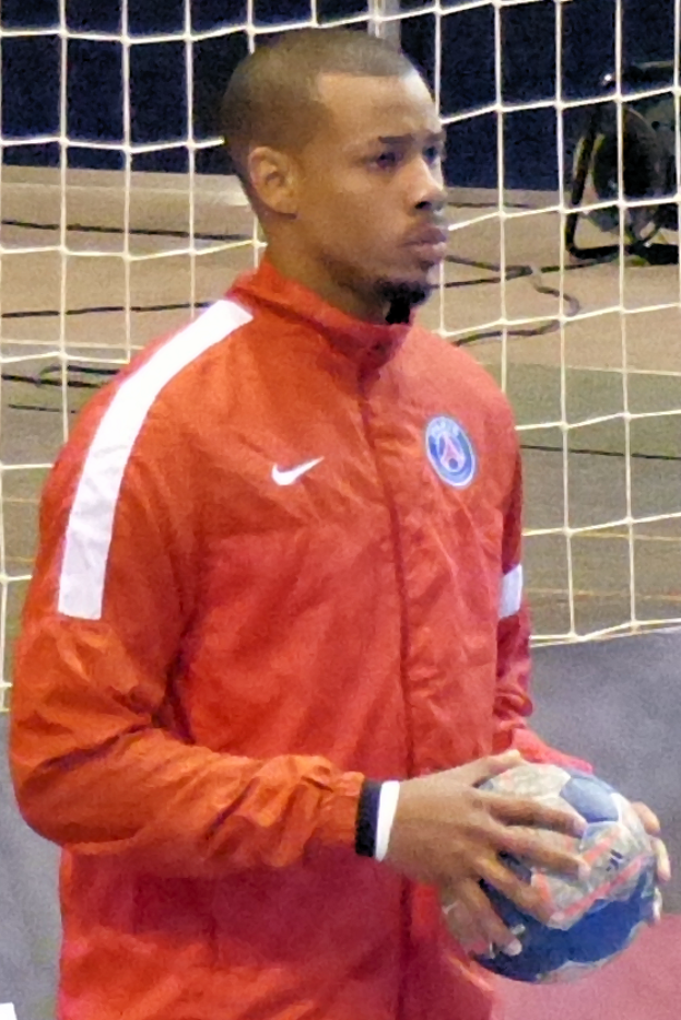 Ein Mann in einem blauen Hemd und Shorts steht vor einem Fußballtor und hält einen Fußball in den Händen, mit einem entschlossenen Gesichtsausdruck und konzentrierten Augen.