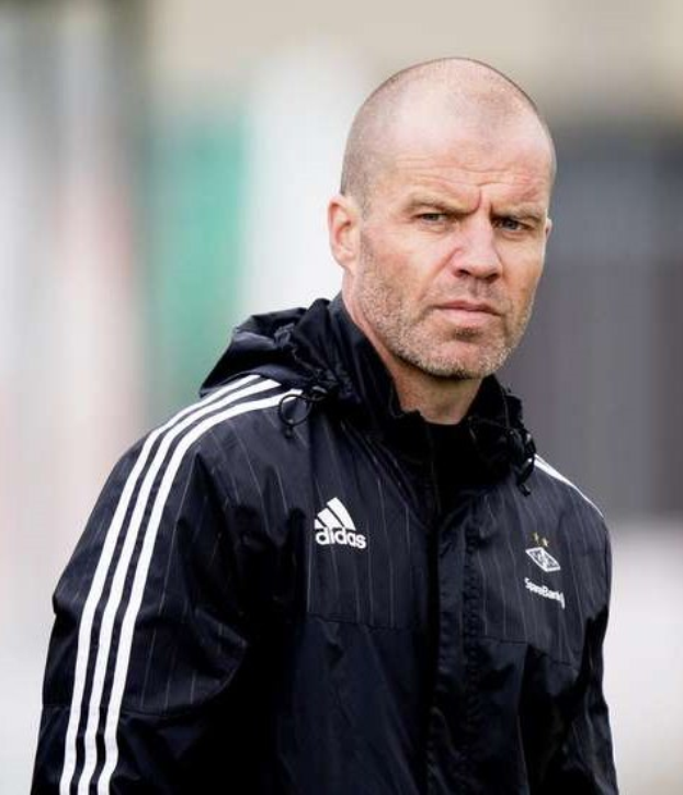 Ein kahlköpfiger Mann in einer schwarzen Jacke steht auf einem Fussballfeld, trägt eine marineblaue Jacke mit weißen Streifen und einen weißen Kragen und hat die Hände verschränkt und einen ernsten Gesichtsausdruck.