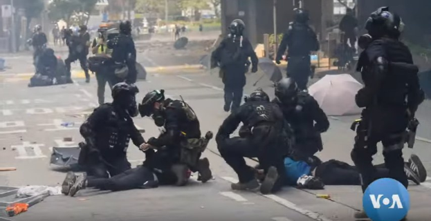 Eine Gruppe von Polizisten in Schutzausrüstung steht um einen verletzten Mann herum, der auf dem Boden liegt, mit Bäumen und Gebäuden im Hintergrund.