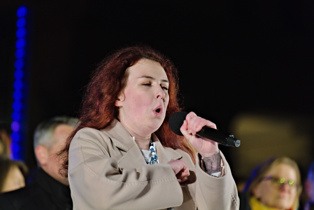 Eine Frau singt in ein Mikrofon auf der Bühne mit zwei Personen dahinter, unter Bühnenlicht mit unscharfem Hintergrund.