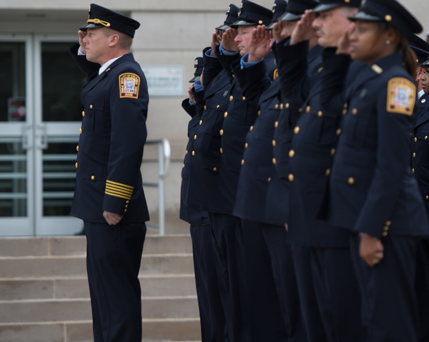 Eine Gruppe von Polizisten in Uniform und mit Mützen steht in Formation und salutiert vor einem Gebäude mit Glasfenstern und Treppen.