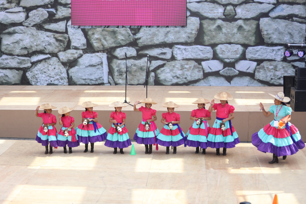 Eine Gruppe junger Mädchen in bunten Kleidern und Hüten tanzt auf einer Bühne mit Verkehrskegeln, Mikrofonen, Lautsprechern, Scheinwerfern und einem Bildschirm mit Text im Hintergrund, während das Publikum unten zuschaut.