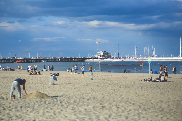 Eine Gruppe von Menschen spielt Volleyball am Strand mit einem Netz, Booten und einer Brücke im Hintergrund bei einem bewölkten Himmel.