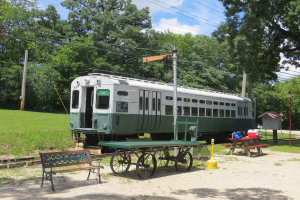 Ein Zug auf Schienen mit einer Bank und einem Tisch in der Nähe, umgeben von Gras, Bäumen und einem Mast mit Drähten unter einem klaren Himmel.