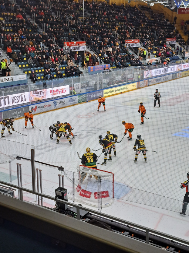Gruppe von Menschen, die Hockey auf einem Eisplatz spielen, umgeben von einem Zaun mit einem Netz in der Mitte, Zuschauern im Hintergrund, Bannern mit Text und Überdachungsbeleuchtung.