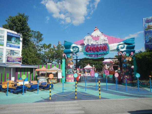 Eine Karussellfahrt am Parkeingang mit einer Straße, einem Zaun, Buden, Banner, Bäumen und einem bewölkten Himmel.