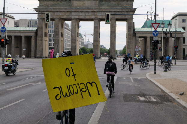 Eine Gruppe von Radfahrern fährt an der Brandenburgertor in Berlin vorbei, einer hält ein gelbes Schild, mit Laternenmasten, Verkehrszeichen, Gebäuden und Bäumen im Hintergrund unter einem klaren blauen Himmel.