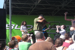 Eine Gruppe von Müsikern, die auf der Bühne mit verschiedenen Instrumenten spielen, während das Öffentlichkeit davor steht und die Musik genießt, mit einem großen schwarzen Lautsprecher links und einem grünen Banner im Hintergrund.