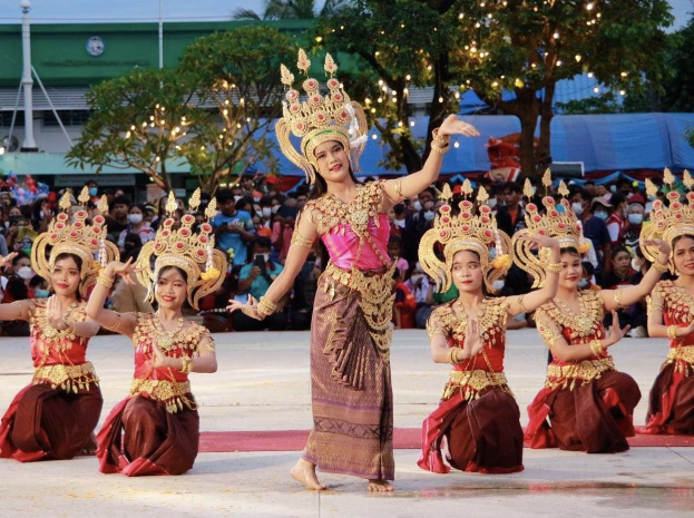 Eine Gruppe von Frauen in traditioneller thailändischer Kleidung, die vor einem Publikum tanzen, mit Bäumen, Lichtern und einem Gebäude im Hintergrund unter einem klaren blauen Himmel.
