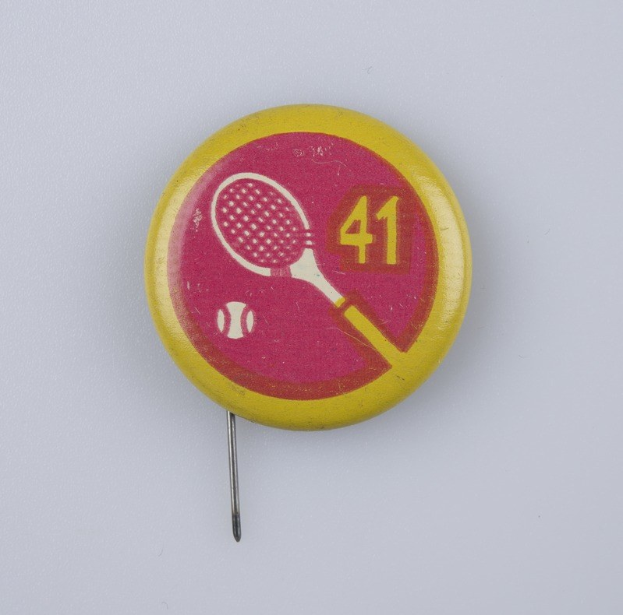 Gelb-rosa Abzeichen mit einem Tennisracket-Design, das mit einer Nadel an einer weißen Oberfläche befestigt ist und Text und Zahlen enthält.