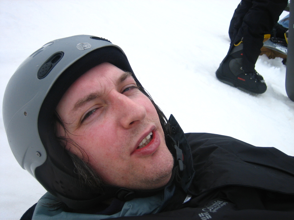 Ein Mann in einem Pullover und Helm steht im Schnee, mit den Beinen und Skiern einer Person in der oberen rechten Ecke.