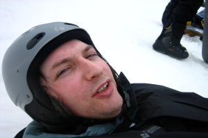 Ein Mann in einem Pullover und Helm steht im Schnee, mit den Beinen und Skiern einer Person in der oberen rechten Ecke.