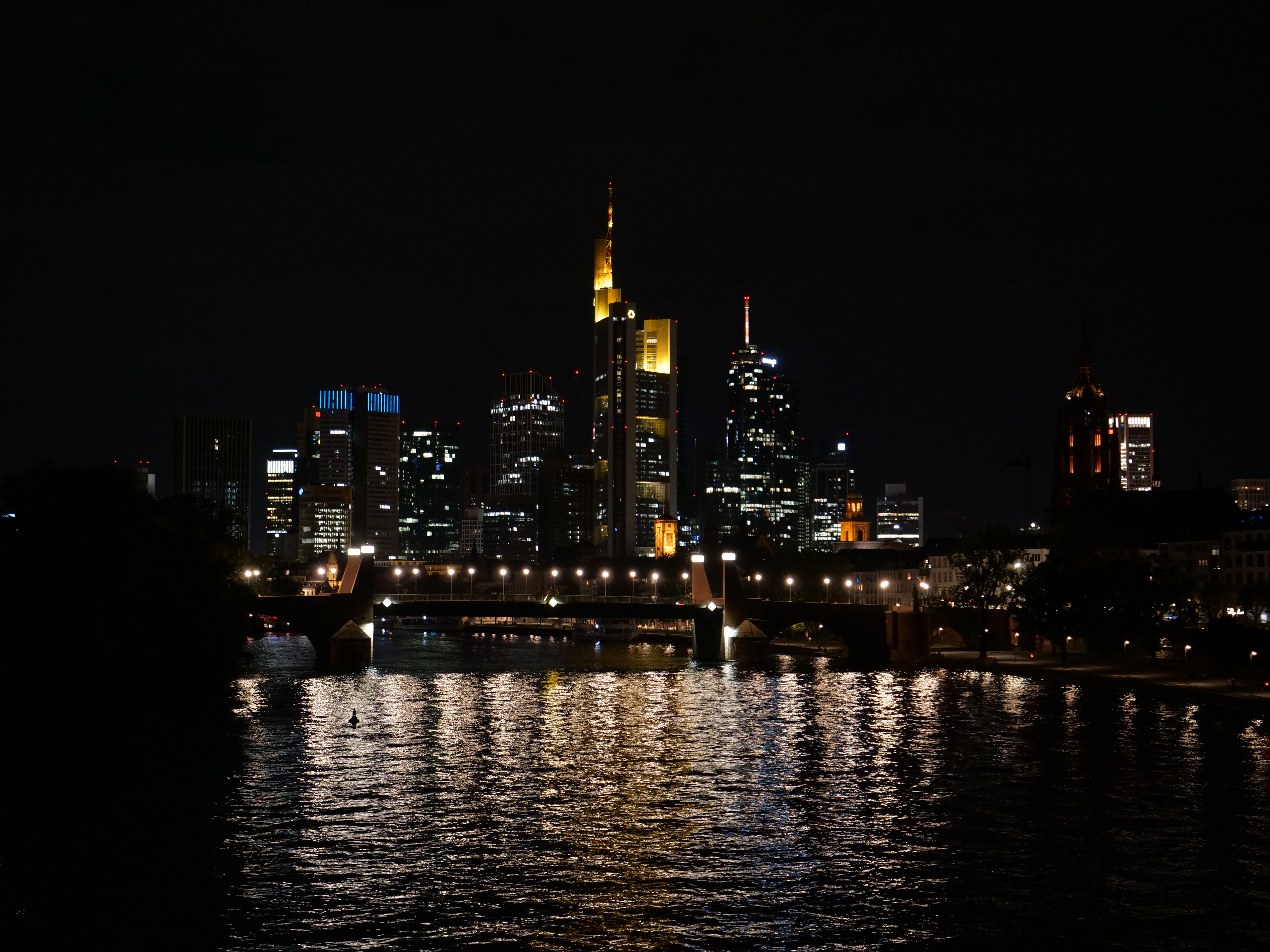 Die Frankfurter Skyline bei Nacht, mit beleuchteten Gebäuden und Sternen im Wasser gespiegelt.