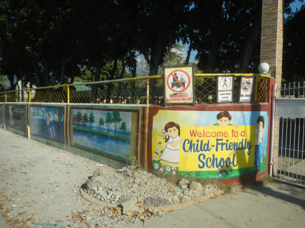 Ein Wandschild mit der Aufschrift "Willkommen in einer kinderfreundlichen Schule" vor einem Metallzaun, Bäumen, Steinen und einem Weg unter einem bewölktem Himmel.