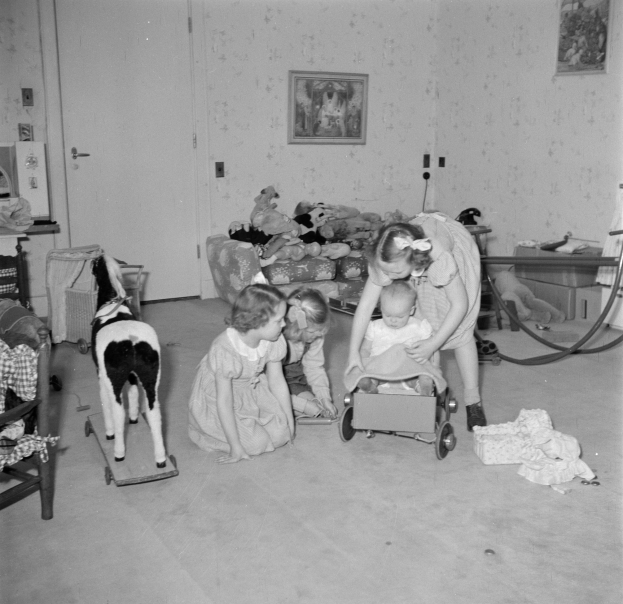 Ein Schwarz-Weiß-Foto von Kindern, die um einen Säugling im Kinderwagen spielen, mit Spielzeugen, einem Hund, Möbeln und Fotorahmen an der Wand.
