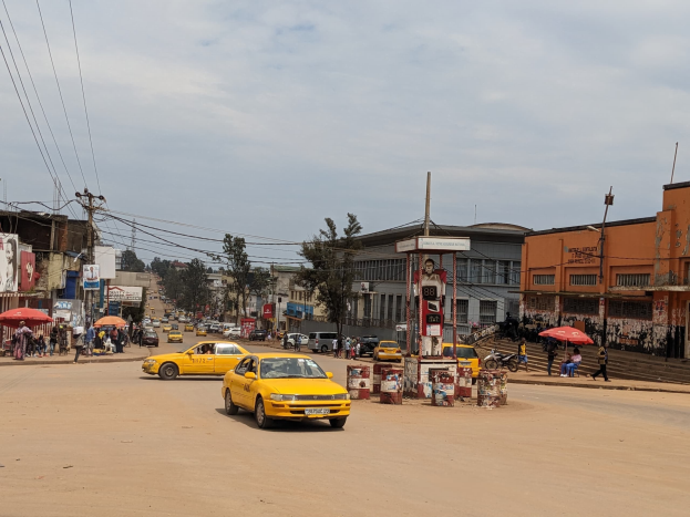 Ein gelbes Taxi fährt eine belebte Stadtstraße mit Gebäuden an den Seiten entlang, während Fußgänger mit Regenschirmen auf dem Gehweg unter bewölktem Himmel gehen.