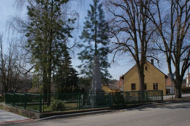 Ein Holocaust-Mahnmal steht in der Mitte einer Straße, umgeben von einem Metallzaun, umgeben von Grün, Häusern, einem Straßenmast und einem bewölkten Himmel.
