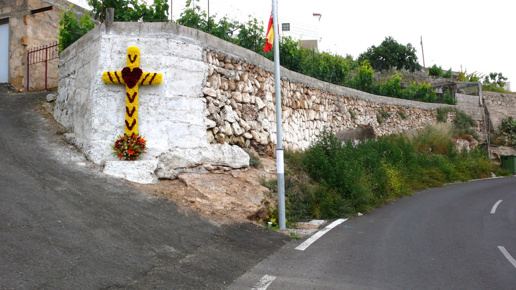 Ein Weg rechts mit Pflanzen, einem Pfahl, einer Fahne, einem christlichen Symbol und Blumen, gesäumt von einer Steinmauer, Bäumen, Drähten und einem Tor links, mit Gebäuden im Hintergrund.