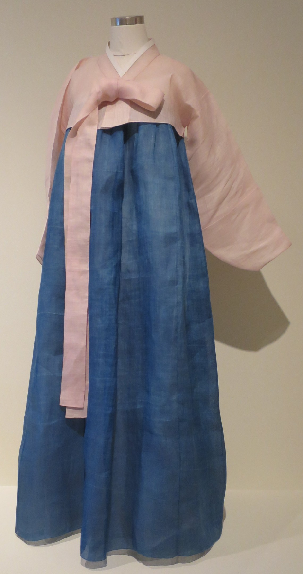 Eine Schaufensterpuppe in einem blauen und pinkfarbenen Hanbok mit komplexen Mustern, die vor einer neutralen Wand in einem Museum ausgestellt ist.