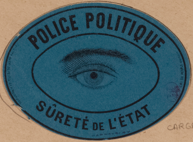 Eine braune Pappkiste mit einem blauen Aufkleber, auf dem "Polizei Politique Sûreté de l'État" steht und ein Auge in der Mitte zu sehen ist.