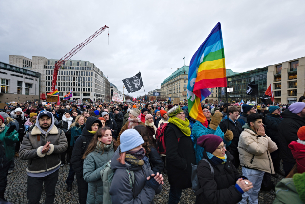 Eine große Gruppe von Menschen mit LGBTQ+-Rechten-Schildern und -Fahnen vor einem Gebäude in Berlin, mit einem Kran und einem bewölkten Himmel im Hintergrund.