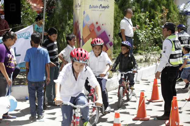 Kinder fahren mit Fahrrädern eine Straße entlang, die mit Verkehrskegeln gesäumt ist, einige tragen Helme, andere stehen in der Nähe; ein Banner, Bäume und Gebäude im Hintergrund.