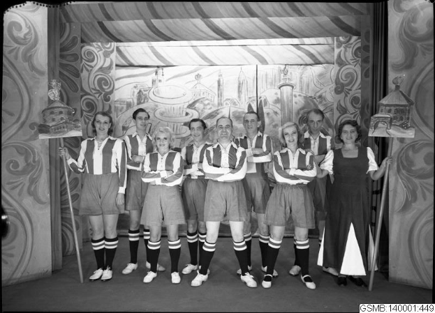 Eine Gruppe strahlender Frauen in 1940er-Kleidung steht auf einer Bühne und hält Stöcke, mit einem Gemälde im Hintergrund und Text unten, der "1940er-Frauenfußballmannschaft" lautet.