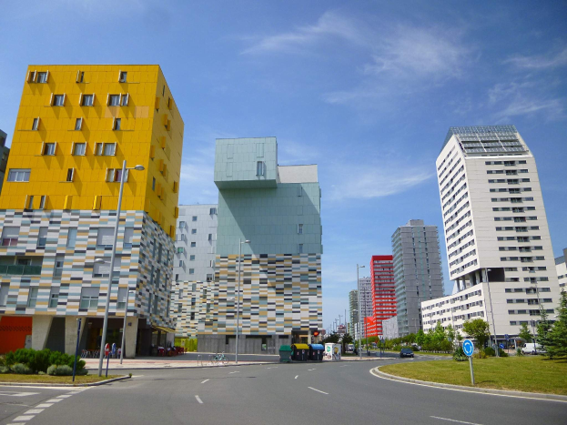 Eine Straße in der Stadt mit hohen Gebäuden im Hintergrund, Straßenmasten, Straßenlaternen, Kraftfahrzeugen, Infotafeln, Büschen, Bäumen und einem bewölkten Himmel, mit Gebäuden der Universität Rotterdam mit einzigartiger Architektur.