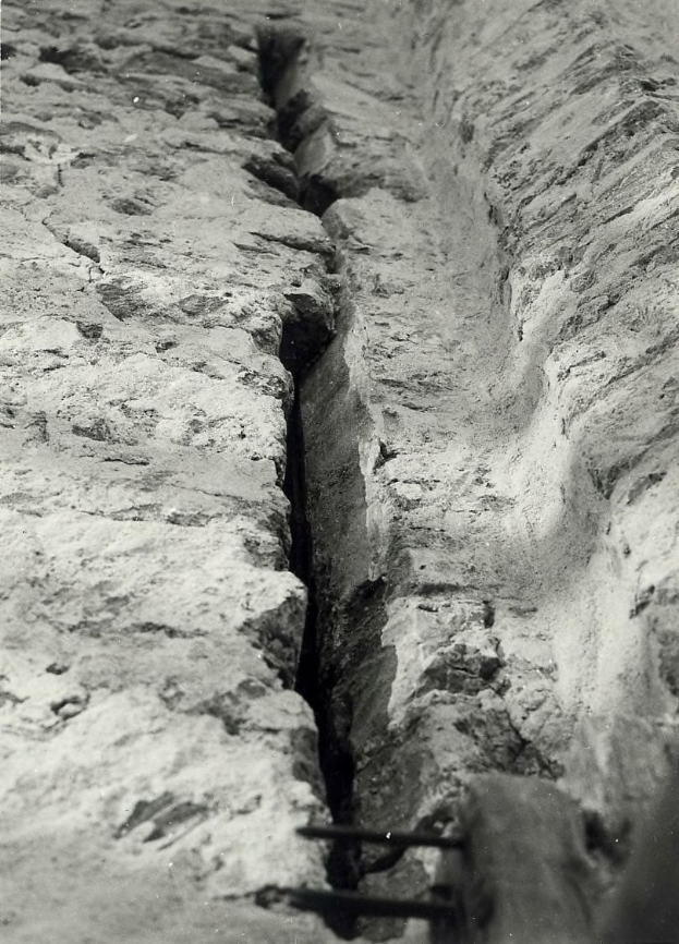 Ein Schwarz-Weiß-Foto eines großeen Risses im Boden, der an einen Entwässerungskanal erinnert, mit einem sichtbaren Wasserzeichen auf der linken Seite.