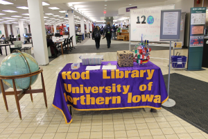 Eine Ausstellung mit einem Tisch, auf dem verschiedene Landesflaggen, eine Box, Papiere und ein Banner liegen, eine Kugel links daneben, ein Anschlagbrett rechts mit ein paar Menschen in der Nähe und mehreren Tischen und Stühlen links.