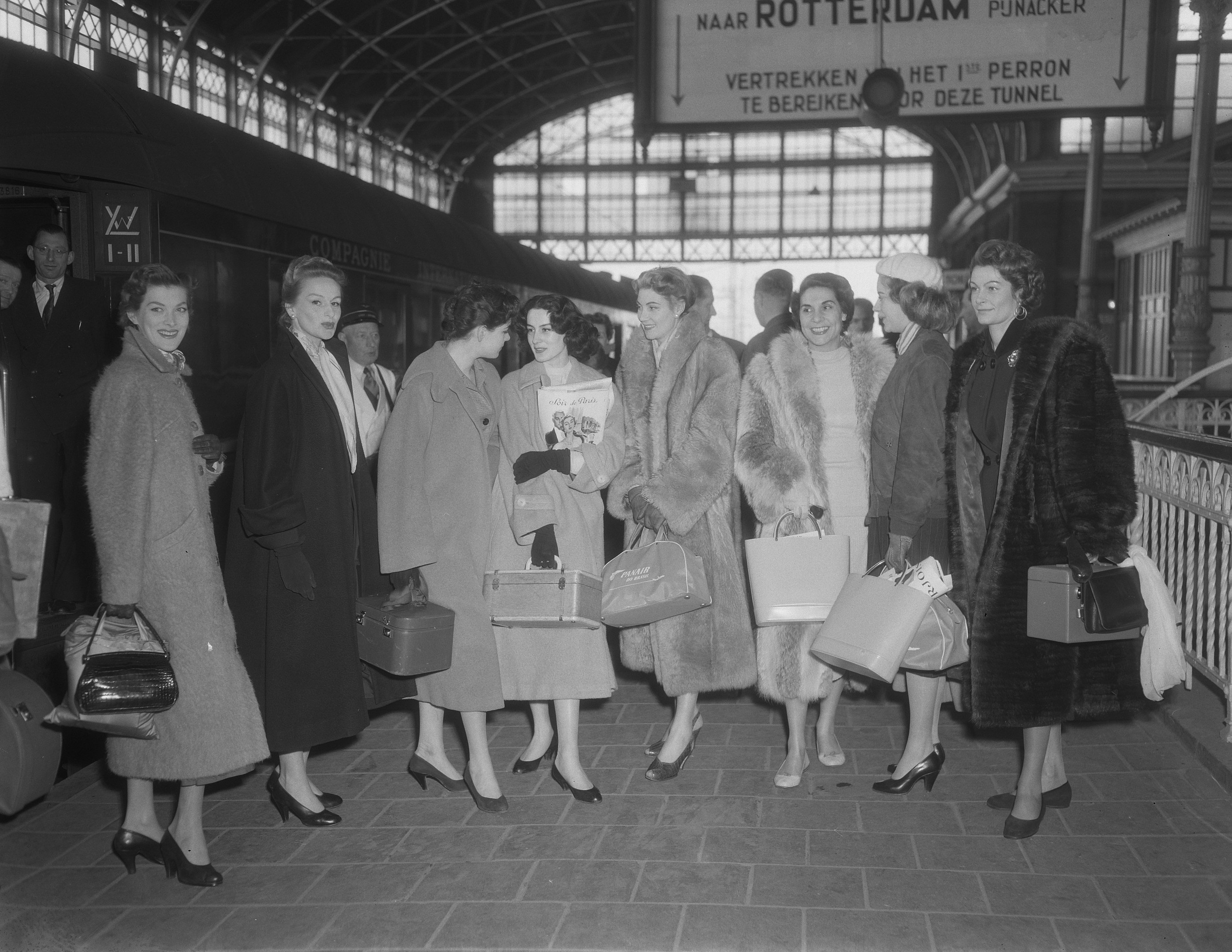 Schwarz-weiß-Bild einer Gruppe von Frauen, die an einem Bahnhof stehen, jede hält eine Tasche, mit einem Zug auf der linken Seite und Geländern und einer Tafel auf der rechten Seite.