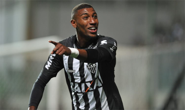 Ein Fußballspieler in einem schwarz-weißen gestreiftem Trikot lächelt, während er ein Tor feiert, wobei der Hintergrund absichtlich unscharf ist.