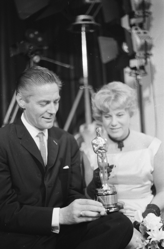 Ein Mann in einem schwarzen Blazer, weißem Hemd und Krawatte hält eine Oscar-Statue und sitzt neben einer Frau, die einen Blumenstrauß hält, beide in formeller Kleidung, mit Lichtern im Hintergrund, auf einem Schwarz-Weiß-Bild.