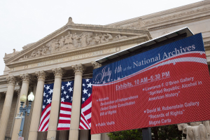 Außenansicht des Nationalarchivs in Washington, DC, mit einem Banner, Fahnen, Laternenmasten, Bäumen und Skulpturen vor einem klaren blauen Himmel.