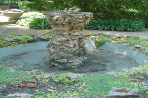 Ein Brunnen umgeben von kleinen Pflanzen mit Bäumen im Hintergrund.