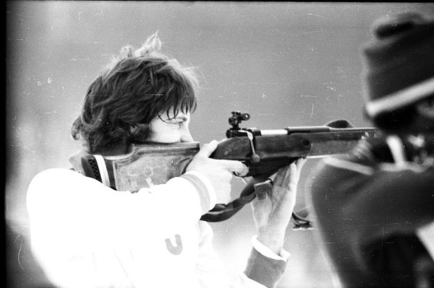 Ein Mann in einem weißen Hemd zielt mit einem Gewehr auf ein Ziel, während rechts neben ihm eine weitere Person steht, dargestellt in Schwarz-Weiß.