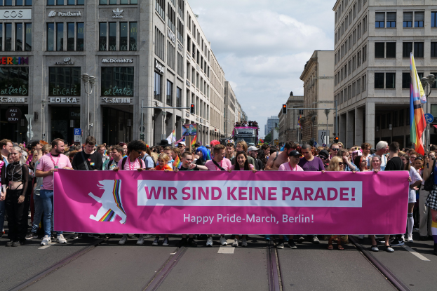 Eine Gruppe von Menschen marschiert mit einem pinken "Happy Pride March"-Schild die Straße in Berlin entlang, mit Gebäuden, Laternenmasten und bewölktem Himmel im Hintergrund.