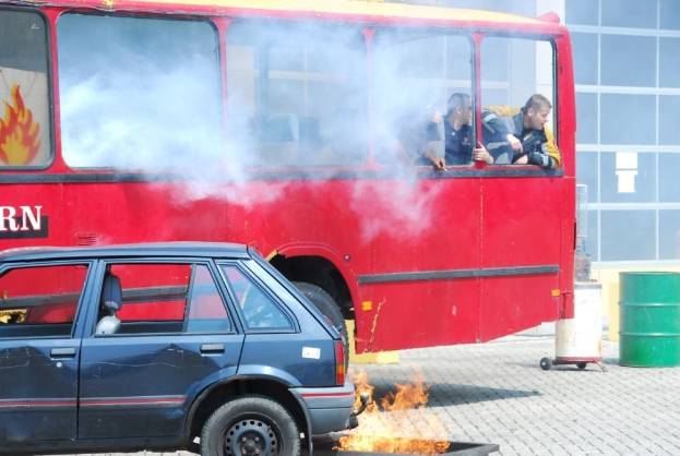 Ein roter Doppeldeckerbus mit Rauch, drei sichtbaren Passagieren, neben einem Auto geparkt, vor einem Gebäude mit Glasfenstern und einem Fass auf der rechten Seite.
