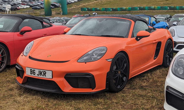 Porsche 718 Boxster GTS auf dem Goodwood Festival of Speed mit anderen Autos, einem Banner, Bäumen und einem klaren blauen Himmel im Hintergrund.