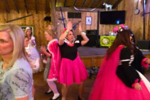 Eine Gruppe von Menschen in Kleidern tanzt in der Mitte eines Raums, während eine andere Gruppe links steht, ein Fernseher, ein Spielzeugtier und verschiedene Gegenstände im Hintergrund zu sehen sind.