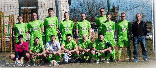 Gruppe von Männern in grünen T-Shirts auf einem Fussballfeld in der Nähe eines Torpfostens mit einem Ball auf dem Boden und verschiedenen Hintergrundelementen wie Bäume, Gebäude und Fahrzeuge.