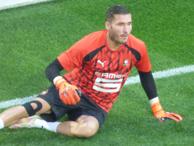 Ein Fußballer in rotem T-Shirt, schwarzen Shorts, weißen Socken und orangefarbenen Baseball-Handschuhen sitzt auf dem Boden mit den Händen auf den Knien.