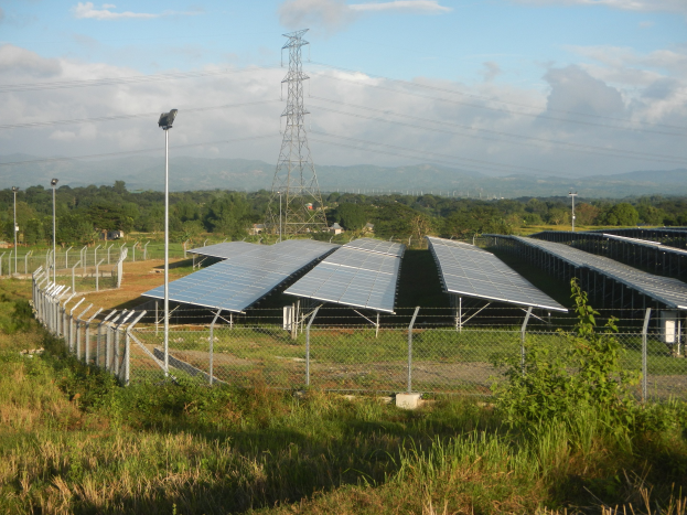 Ein Feld von Solarpanels hinter einem Zaun, mit Gras, Pflanzen und Bäumen und einem Übertragungsturm mit Drähten im bewölkten Himmelhintergrund.