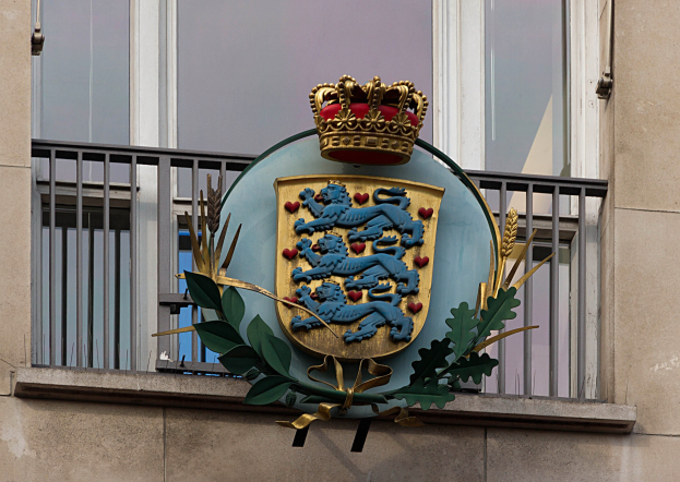 Wappen der britischen Königsfamilie auf einem Gebäudeäußeren angezeigt, begleitet von einer Krone und einem Blatt-Logo, mit Fenstern und Metallgittern im Hintergrund.