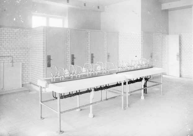 Schwarz-weiß-Foto einer öffentlichen Toilette mit einer Reihe von Waschbecken, Wasserhähnen, Wänden, Türen, Fenstern und einem Boden, mit verschiedenen Gegenständen darauf verstreut.