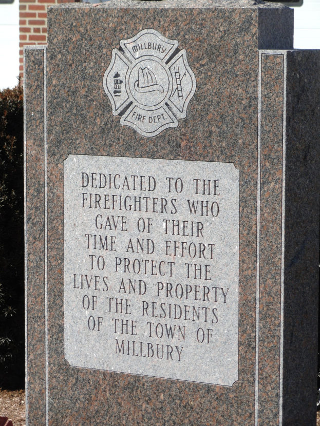 Denkmalstein mit einer Plakette für Feuerwehrleute in Millbury, vor einem Backsteingebäude inmitten von Grünanlagen.